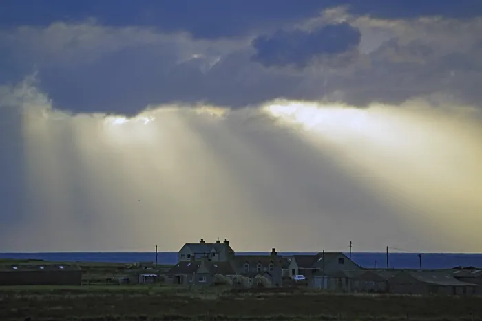 Gerahmter Druck : Sonnensstrahlen brechen durch dramatische Wolken über Häuser am Meer.