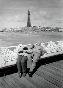 Drucke von Ein älteres Paar liegt sich auf die von Eisen verzierte Sitzplätze am Blackpools Pier ausruhen. Die Strand und das Blackpools Turm sind im Hintergrund zu sehen. Datum: ca. 1970 Schlafzimmer Drucke: Schlafen auf der Blackpools Promenade