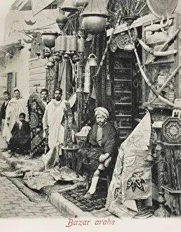 Drucke von Tunesien - ein arabischer Händler sitzt im Laden und umgeben von seinen Waren (nichts ist ihm fremd) einschließlich Vorhängen , Teppiche , Topflese , eine gestopfte Babykrokodil, Schwerter, Gewehre Tunesien Drucke: Tunesien - arabisches Souk