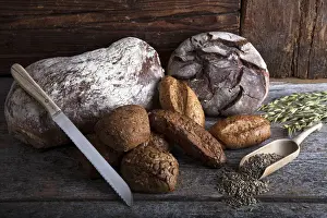 Brotlaibe und Hefebrötchen sowie eine Brotechnik aus Stahl auf einem gerodeten Holztafel mit Roggenkörnern