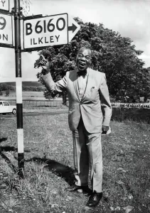 Drucke von Amerikanischer Jazzmusiker Louis Armstrong vor einem Schild im Yorkshire, der den Weg zu Ilkley zeigt Juni 1968 P003769 0000bis0099 Drucke: Amerikanischer Jazzmusiker Louis Armstrong vor einem Schild im Yorkshire