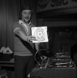 Drucke von Die Tänzer des Wigan Casinos aus dem Jahr 1975 halten einen DJ namens Russ Winstanley fest, der eine Singleplatte hält und neben seinen Plattenständen steht 0000bis0099 Drucke: Tänzer des Wigan-Casino aus dem Jahr 1975 halten den nördlichen Soul-DJ Russ Winstanley eine Single auf