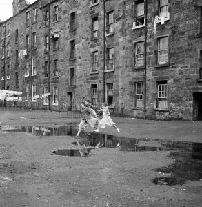 Drucke von Drei junge Freunde springen über einen Pfuhl vor einem Wohnblock von Govan in Glasgow im September 1956 Vereinigtes Königreich Drucke: Drei junge Freunde springen über einen Pfuhl vor einem Wohnblock aus der Gegend von Govan in Glasgow