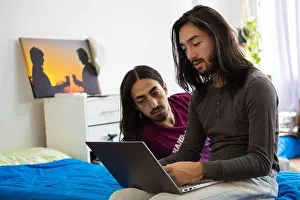 Zwillingsbrüder liegen im Bett und schauen auf den Computer, 2 langehaarige Männer schauen auf den Computer, liegen auf dem Bett.