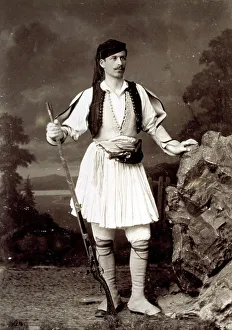 Ein junger Griechen in traditioneller Kleidung mit Minirock und niedriger Jacke, auf dem Rücken eine fiktive Felsplatte tragend und bewaffnet mit einer Pistole.