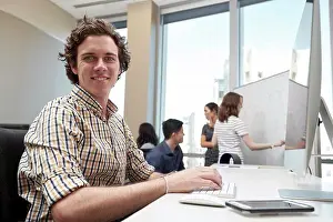 Businessman In Office Drucke: Young man using computer in office looking towards camera