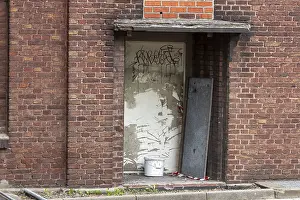 Wandmalereien Drucke: Verlassener Eingang mit Graffiti auf einer Ziegelmauer und Anzeichen von Verfall.
