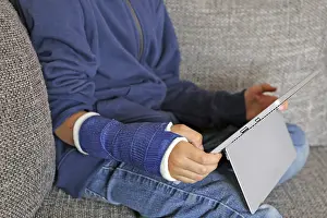 Young Boy On Sofa Drucke: Boy with arm in plaster sits on the sofa with a tablet