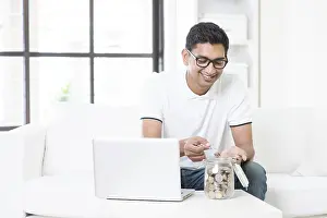 Man Relaxing At Home Drucke: Indian guy using internet computer and counting coins at home