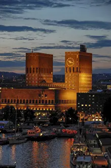 Drucke von Ein großes Gebäude mit zwei Türmen und einem Uhrturm, beleuchtet am Abend mit Booten im Wasser, Oslo, Norwegen Oslo Drucke: Ein großes Gebäude mit zwei Türmen und einer Uhr.