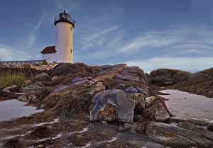 Lighthouse At Sunset Drucke: Lighthouse surrounded by rocks at sunset