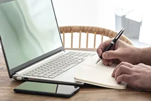 Man Working From Home Drucke: man taking notes on paper while using laptop computer