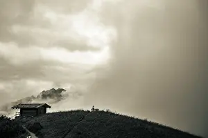 Hiking In The Alps Drucke: Mountain hut with bench and climber and dramatic clouds in the background