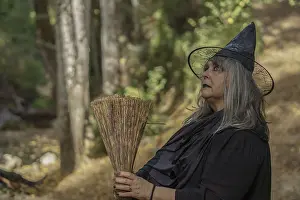 Drucke von Alte Hexe in Schwarz, bekleidet in Schwarz, gesehen im Profil mit ihrem Besen und einem Wald im Hintergrund Schwarze Hexe Drucke: Alte Hexe in schwarz, bekleidet in schwarz.