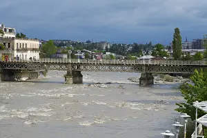 Drucke von Ein ländlicher Brücke über einen Fluss mit einem urbanen Hintergrund unter wolkenverhangenem Himmel, Weiße Brücke über den Rioni-Fluss, Kutaisi, Imeretien-Region, Georgien Kutaisi Drucke: Ein rustikaler Brücke über einem Fluss mit einer städtischen Kulisse unter einem bewölkten Himmel