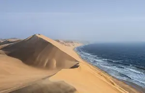 Drucke von Ein atemberaubendes Bild der Namibwüste, wo sich die endlosen Dünenwellen mit den Wellen des Atlantiks treffen lassen Kulturerbestätten Drucke: Sanddünen mit Wellenmustern und See bei Abendlicht
