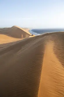 Drucke von Ein atemberaubendes Bild des Namib-Dünenlandschafts, beleuchtet von der Abendsonne Kulturerbestätten Drucke: Dünenlandschaft mit Wellenmustern und Meeresblick bei Abendlicht