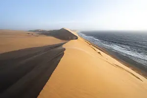 Drucke von Dünen mit Wellenmustern und Meer im Abendlicht, Wind bläst Sand über die Dünen, Atlantikküste und sandiger Wüstensand mit einem Meer aus Dünen, beeindruckende Wüstentopographie bei Sonnenuntergang Kulturerbestätten Drucke: Dünen mit Wellenmustern und Meer am Abendlicht.