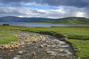 Cloudy Sky With Mountains Drucke: A small river flows through green fields under a cloudy sky with mountains in the background