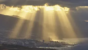 Canary Islands Scenery Drucke: Sun rays break through dramatic clouds over a coastal town
