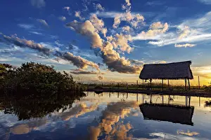 Evening Sun Reflection Drucke: A thatched hut reflected in the water at sunset with dramatic cloud formations