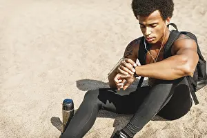 Single Person View Drucke: Young man sitting on a sand and looking at his activity tracker